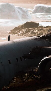 In a desolate area, a crashed plane rests among icy formations and rocks near the ocean. The sunset casts a warm light over the scene, highlighting the wreckage.