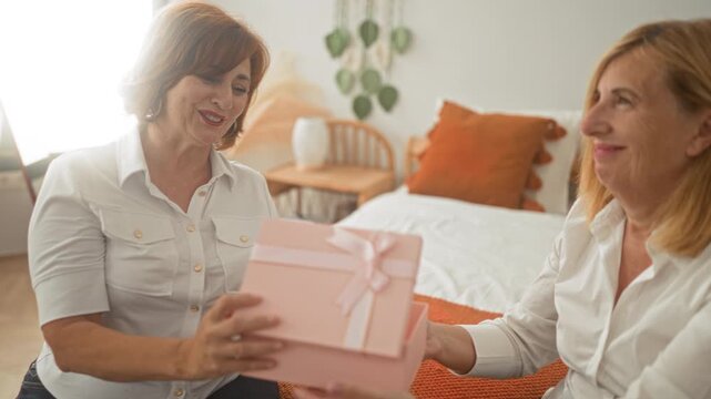 Woman handing a pink gift box to another woman on bed in bedroom while smiling and later hugging in warm domestic scene; affection joy.