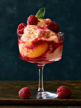 gourmet peach melba dessert in a stemware glass featuring creamy vanilla ice cream, juicy peach slice, fresh raspberries, mint leaf, and sweet red berry syrup topping served on a rustic dark table