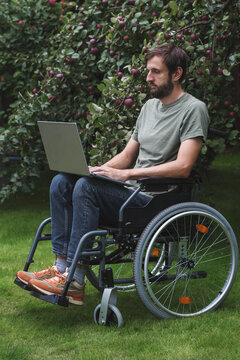 Young bearded man using laptop while sitting in wheelchair outdoors near apple tree, illustrating accessible technology, remote work, independence, inclusion, disability, modern lifestyle vertical
