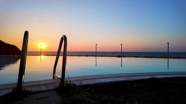 Sunset just above the horizon, over a pool with crystal clear, light blue water, an infinity pool overlooking the calm sea