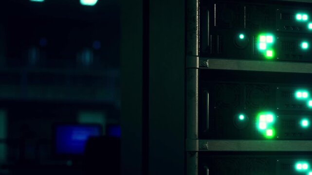 Several green lights on a server rack indicate active devices in a data center during nighttime. Computer monitors can be seen in the background, adding to the high tech environment.