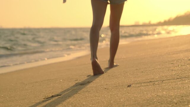 Happy beautiful woman walking on sand beach enjoying sunset day of leisure and freedom, close up of barefoot legs walking along seashore with gentle waves under golden sky.