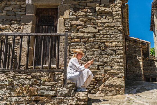 Woman using smartphone while sitting on stone steps in medieval village of Rupit, Catalonia, Spain, checking travel information