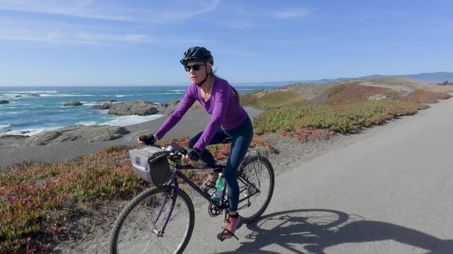 Mature woman rides her bike on a paved trail in Fort Bragg near Mendocino California next to the ocean under a clear sky.