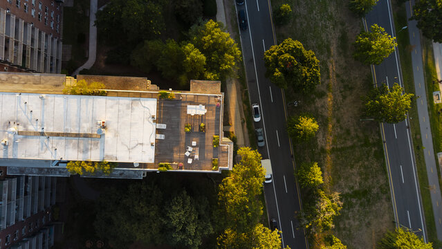 Aerial view of a residential building rooftop with a wooden deck and patio furniture, adjacent to a tree-lined road with cars in Boston, Massachusetts, United States.