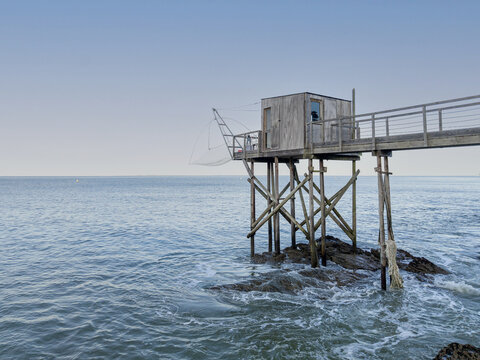 Aerial view of a traditional wooden fishing hut on stilts with a large net extending over the calm sea and rocky coastline in Pornic, Pays de la Loire, France.