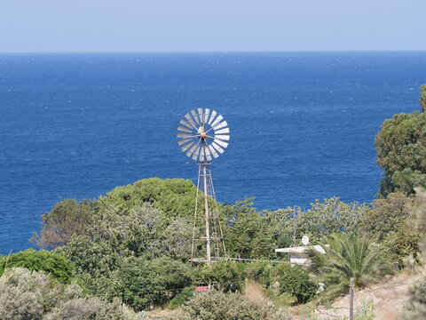 traditional steel water-pumping windmill stands among green trees on a hillside, overlooking the deep blue sea under a clear sky.