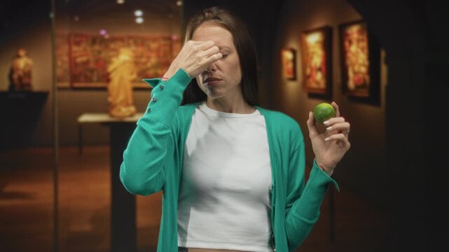 Woman pinches nose with fingers while holding a lime near her face in an indoor art gallery building lit by spotlights and framed paintings; disgust.