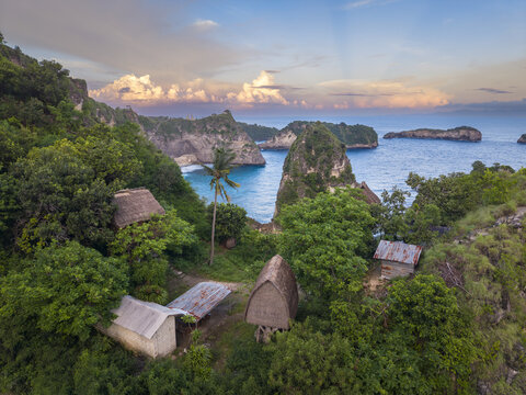Aerial view of traditional thatched-roof huts nestled among lush green vegetation overlooking the rocky cliffs and blue ocean of Diamond Beach in Nusa Penida, Bali, Indonesia.