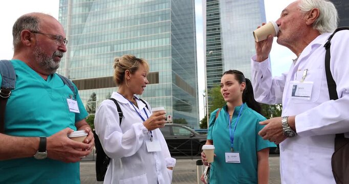 Group of doctors and nurses in uniforms enjoying a coffee break together, talking and relaxing outside a modern building, representing healthcare professional camaraderie and work life balance