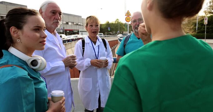 Multiracial group of doctors and nurses wearing uniforms taking a coffee break outside the hospital building. Medical staff members relaxing and talking during their work shift outdoors
