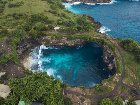 Aerial view of Broken Beach featuring a natural rock archway over turquoise ocean waters, surrounded by lush green cliffs and walking paths in Nusa Penida, Bali, Indonesia.