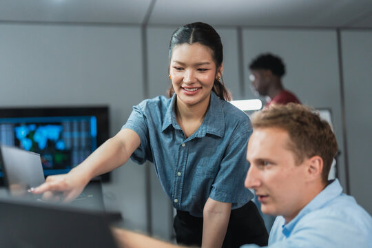 Young developer points at a dashboard on his monitor while an Asian colleague leans in to look, discussing analytics results together in a modern software development office.