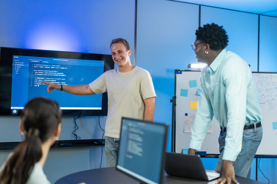 Smiling developer points at code on screen while colleague listens beside whiteboard with sticky notes, software team in sprint planning meeting in modern blue lit office