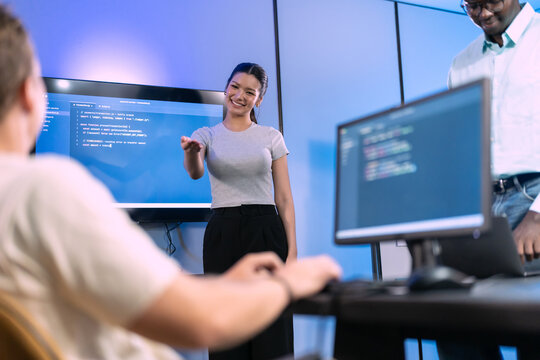 Young woman leads daily standup while colleagues listen, software development team gathered around monitors and wall screen with code in blue lit modern workspace
