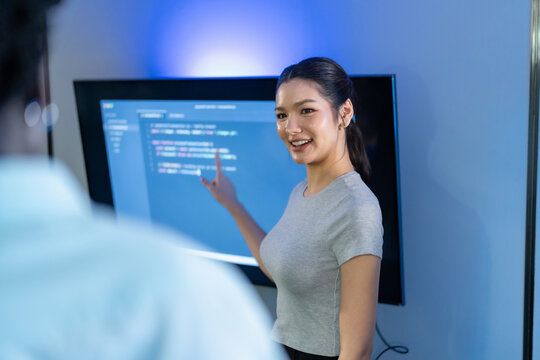 Smiling young woman points at lines of code displayed on screen while explaining software project to colleague, female developer leading technical session in modern office