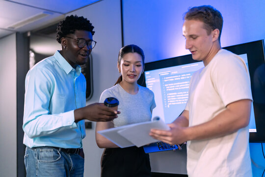 Three colleagues stand together reviewing printed documents on clipboard, multiethnic business team discussing project details in blue lit office with screen behind