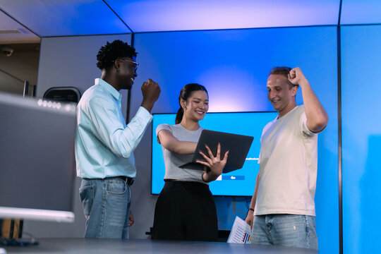 Three colleagues celebrate good news on laptop screen with raised fists and smiles, multiethnic startup team sharing project success in blue lit modern office