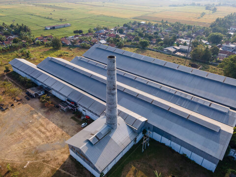 Aerial view of a large industrial factory with a tall brick chimney, surrounded by lush green rice fields and residential houses in Banyumas Regency, Central Java, Indonesia.