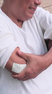 elderly asian man suffering from elbow tendonitis, having pain relieving patch on his elbow, clenching his jaw in pain on a park bench during daytime, vertical video