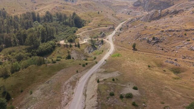 Drone Aerial View Flying Over Winding Gravel Road Through Mountain Valley with Forest and Arid Slopes on Visocica