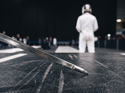 close up of the tip of a fencer foil on the strip in a sports hall, with a fencer in a white protective suit and mask standing in the background