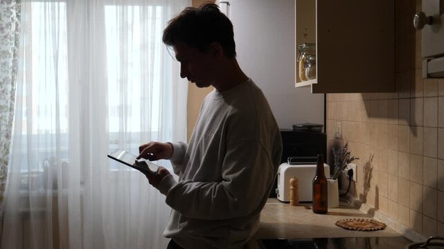 Vertical video man standing in a modern kitchen, viewing a digital recipe on a tablet screen, embodying digital lifestyle and smart home technology while preparing a meal