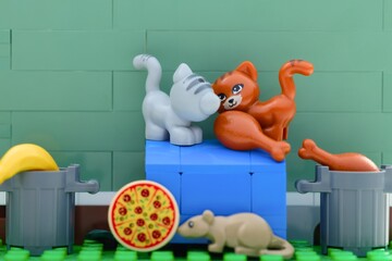 Naklejka premium Tambov, Russian Federation - August 03, 2026 Two Lego cats and a mouse minifigures searching for food like pizza and chicken near trash cans.