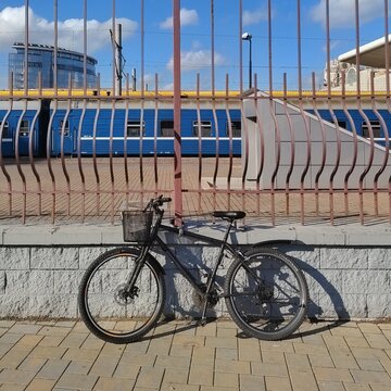 Black urban bicycle with a front basket parked near a metal fence. Blue passenger train and city building in the background