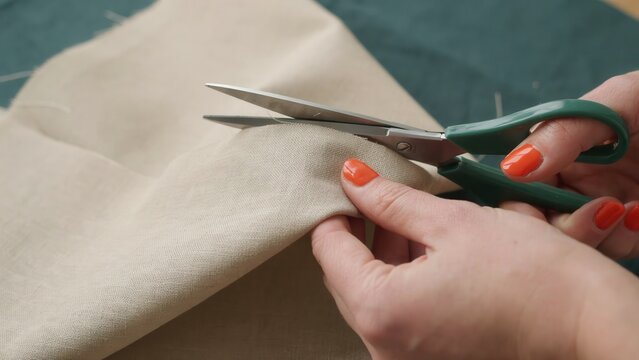 A woman's hands use green scissors to cut beige linen fabric on a green surface. This represents the sewing and tailoring craft process.