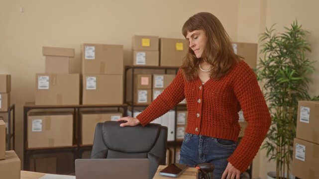 Woman with hand on head and other hand on hip beside stacked cardboard shipping boxes at a packing desk in a building; small business concern.
