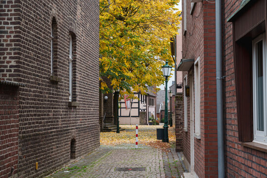 A narrow cobbled alley winds between brick buildings beneath glowing autumn foliage in Zons, Germany