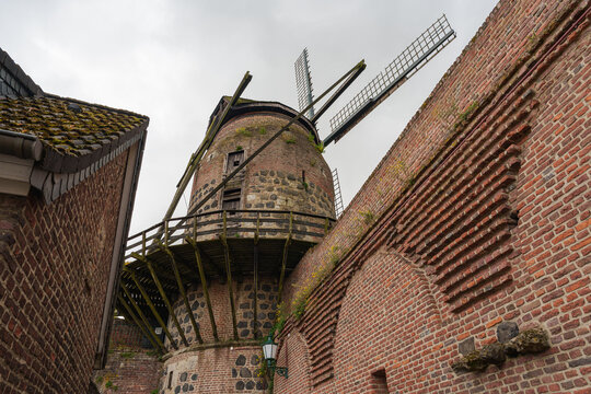 A historic windmill rises above stone arches and medieval walls beneath a cloudy sky in Zons, Germany