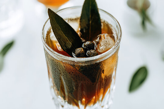 Dark botanical cocktail in crystal glass with sage leaf and juniper berry garnish, condensation and ice against bright bar background