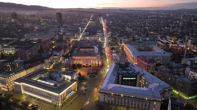 Aerial footage of Sofia, Bulgaria at dusk, showing the illuminated city center, busy boulevards, historic buildings, urban skyline and Vitosha Mountain in the background.