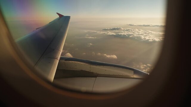 Airplane Wing Over a Cloudscape with Sun Rays Piercing Through Clouds at Sunset. View from Airplane Window of Wing Above Clouds Illuminated by Golden Sunset