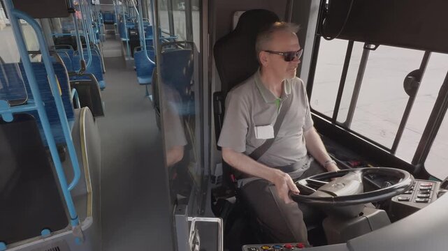 Male bus driver in gray shirt and sunglasses sits at the wheel, focused on driving. The interior of the bus shows empty blue seats and large windows, indicating an urban public transport environment