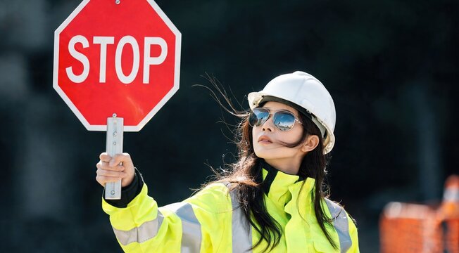 A young Asian woman wearing a hard hat and sunglasses holds a stop sign. She is dressed in a bright yellow safety jacket, standing at a construction site.