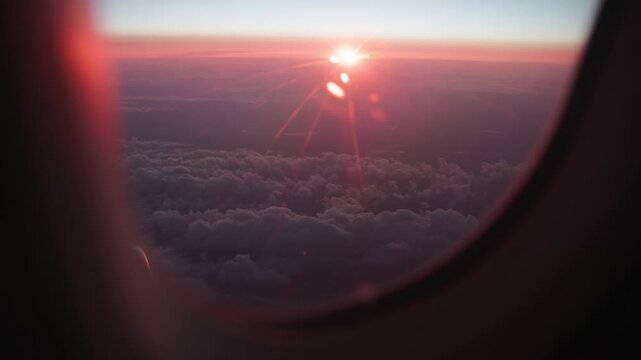 Airplane Window View of Purple Clouds and Red Sunset with Sun Rays. Dramatic Red Sunset Above Purple Clouds with Sunbeams Seen from an Airplane Window
