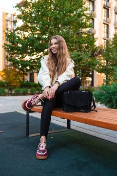 woman relaxing on wooden bench in urban park with trees and buildings in background, side view, casual and relaxed mood, beige sneakers, black bag, leather jacket, fashion, lifestyle, urban.