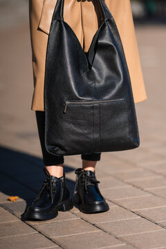 person with large black leather bag standing on urban sidewalk in casual setting with tan coat and black clothing, close-up of lower body and bag in fashion street style.