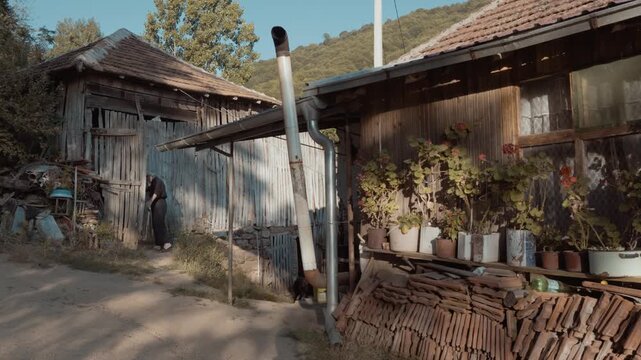 An elderly villager opening a traditional wooden gate door and entering a rustic yard surrounded by old wooden buildings with potted flowers on Stara Planina, Serbia. Authentic rural life.