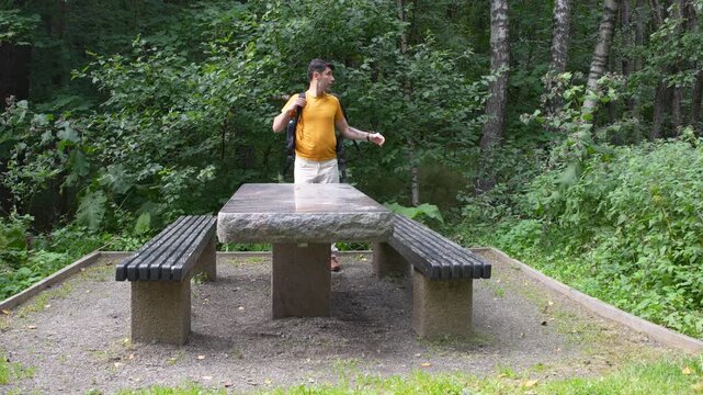Young woman sitting on a wooden bench in a peaceful forest park, wearing a yellow T-shirt and white jeans. Natural outdoor lifestyle scene surrounded by green trees and summer vegetation.