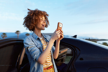 Naklejka premium Cheerful woman outdoors, smiling while holding a phone, beside a car, sunny coastal scene, denim shirt, striped top, relaxed lifestyle, natural colors, soft breeze, warm light, palm trees.
