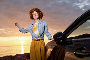 Naklejka premium Young woman with curly hair smiles beside a car at sunset, wearing a denim jacket over a striped top and a yellow skirt, embodying a carefree lifestyle and warm golden light.