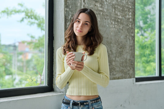 Young attractive woman holding coffee mug standing near window looking at camera