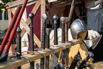 Naklejka premium Row of medieval swords displayed on wooden stand with steel knight helmet at historical festival. Heritage scene, medieval history, reenactment, armor, weapons, cultural tourism