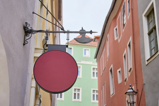 Blank round sign hanging from decorative metal bracket between colorful buildings