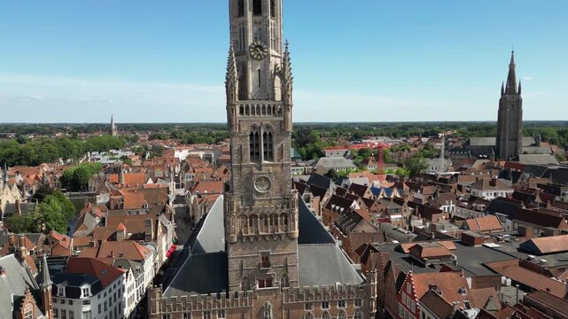Drone camera moving upwards in a pedestal shot, revealing the historic medieval Belfry bell tower and the panoramic urban landscape of Bruges on a sunny summer day.